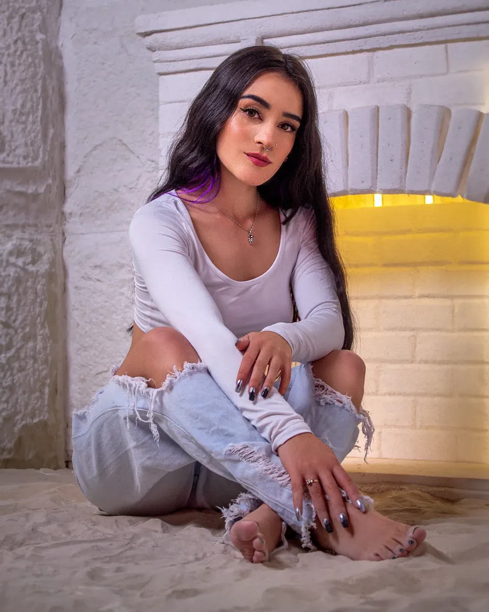 Modelo sentada en la arena frente a una chimenea con luz amarilla, con top blanco y jean.
