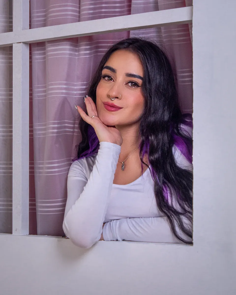 Modelo apoyada en una ventana blanca con cortina lila, con la mano en la mejilla.