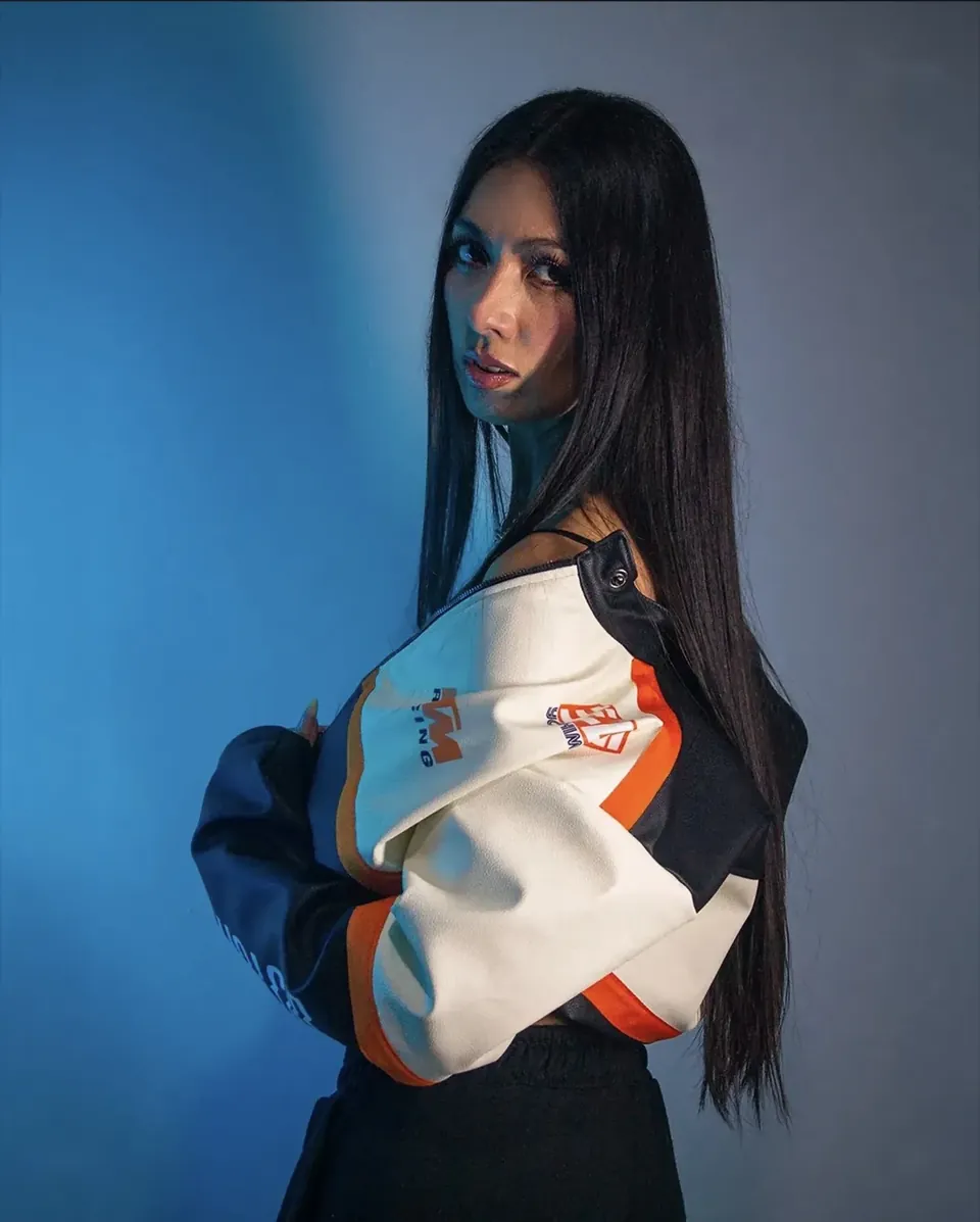 Modelo de cabello largo y negro, de perfil, con chaqueta blanca, negra y naranja sobre un fondo de luz azul.
