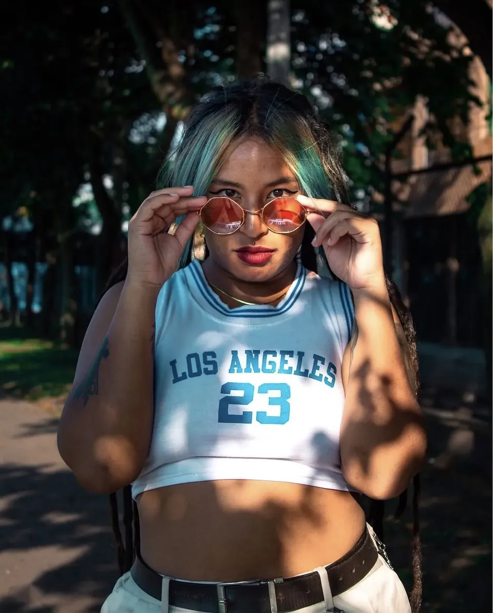 Modelo con mechas verdes y gafas rosadas, camiseta blanca «Los Angeles 23», entre sol y sombras de árboles.